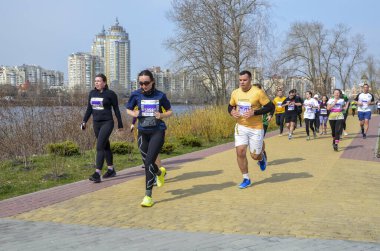 Nehir kenarındaki güneşli şehir parkında çeşitli koşucular yarışırken, arka planda şehir silüeti yer alıyor. Parlak giysiler içindeki enerjik sporcular canlı yaratıyor, yarış atmosferi yaratıyorlar.
