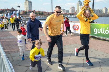 Nehir kenarındaki bir gezinti alanında, gönüllülerin ve destekçilerin rehberliğinde çocuklar ve yetişkinlerin önlük giydiği canlı bir yardım etkinliği düzenleniyor.