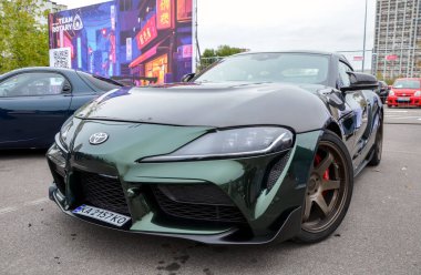 Benzersiz metalik koyu yeşil boya kaplamalı, çarpıcı özelleştirilmiş Toyota Supra A90 nesli açık hava otomobil etkinliklerinde düşük ve ön açılı bir perspektiften görüntülenir.