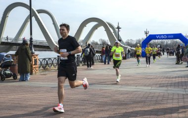 Koşucular, nehrin kenarındaki tuğla bir gezinti güvertesi boyunca ilerlemeye devam ediyor. Uzakta mavi bir kavis var ve seyirciler korkuluklardan izliyor.