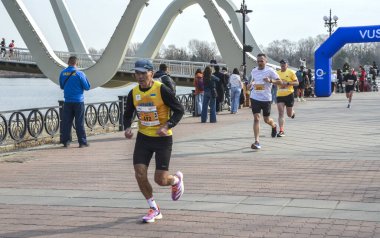 Koşucular, nehrin kenarındaki tuğla bir gezinti güvertesi boyunca ilerlemeye devam ediyor. Uzakta mavi bir kavis var ve seyirciler korkuluklardan izliyor.