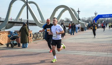 Koşucular, nehrin kenarındaki tuğla bir gezinti güvertesi boyunca ilerlemeye devam ediyor. Uzakta mavi bir kavis var ve seyirciler korkuluklardan izliyor.