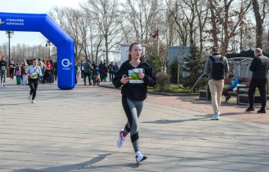 Genç bir kadın gündüz açık hava yarışında kameraya doğru koşuyor. Seyirciler ve diğer katılımcılar mavi kemerin altında kuyruk oluşturuyorlar.