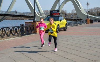 Parlak atletizm kıyafetleri içinde iki genç kız arka planda modern köprü ile rıhtım boyunca koşuyorlar enerji yarışması ve aile dostu spor gösterileri yapıyorlar.