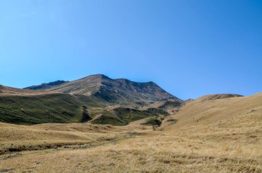 Berrak, parlak mavi gökyüzünün altında yuvarlanan dağların görüntüsü. Ön taraftaki yamaçlar kuru, tawny otlarla kaplıdır, yamaçlardan yukarı doğru koyu yeşil çalılıklara dönüşürler. Georgia