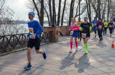 Bir grup koşucu nehir kıyısındaki bir kaldırım boyunca yarışıyor. Katılımcılar finişe doğru ilerlerken güneş ışığı uzun gölgeler oluşturuyor, fitness ve takım çalışması sergiliyor