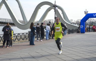 Erkek koşucu, seyircilerin izlediği nehir kenarındaki bir gezinti alanında yarışıyor. Kavisli köprü ve mavi şişme kemer canlı yarış pistini ve şehir atmosferini tanımlar.