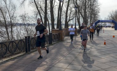 Bir grup koşucu nehir kıyısındaki bir kaldırım boyunca yarışıyor. Katılımcılar finişe doğru ilerlerken güneş ışığı uzun gölgeler oluşturuyor, fitness ve takım çalışması sergiliyor