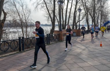 Bir grup koşucu nehir kıyısındaki bir kaldırım boyunca yarışıyor. Katılımcılar finişe doğru ilerlerken güneş ışığı uzun gölgeler oluşturuyor, fitness ve takım çalışması sergiliyor