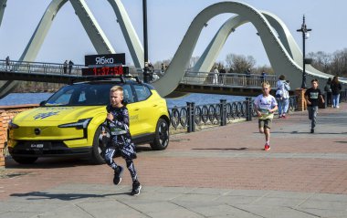 Genç koşucular, çarpıcı bir kemer köprüsünün yanındaki nehir kenarındaki gezinti alanı boyunca yarışıyorlar. Güneşli bir günde çocukların finişe doğru koşmasını sıradan bir kalabalık izliyor.