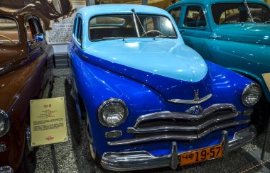 İki ton mavi GAZ-M20 Pobeda sedan, ikonik bir Sovyet dönemi arabası, kendine özgü ponton tarzı kaportası, büyük krom ızgara ve yuvarlak lambalar sergiliyor.