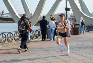 Dişi sporcu modern bir köprünün altındaki nehir kenarında koşuyor. Parlak atletizm teçhizatı enerji ve rekabet taşır