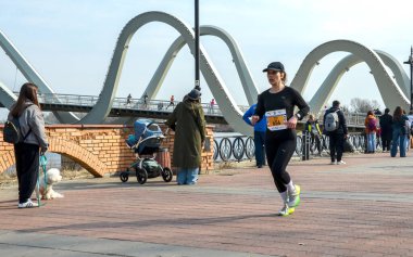 Dişi sporcu modern bir köprünün altındaki nehir kenarında koşuyor. Parlak atletizm teçhizatı enerji ve rekabet taşır