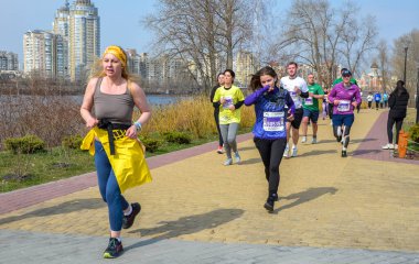 Bir grup koşucu nehir kenarındaki tuğlalı kaldırım boyunca koşuyor ve arkasında modern şehir silüeti bulunuyor. Sahne fitness, takım çalışması ve açık hava kentsel aktiviteleri taşır