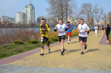 Bir grup koşucu nehir kenarındaki tuğlalı kaldırım boyunca koşuyor ve arkasında modern şehir silüeti bulunuyor. Sahne fitness, takım çalışması ve açık hava kentsel aktiviteleri taşır
