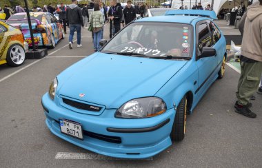 Saldırgan bir şekilde modifiye edilmiş mavi Honda Civic EK hatchback, öne çıkan arka kanat, altın çoklu konuşmalı alaşımlı tekerlekler, kırmızı fren ayarlı ve süspansiyonları indirilmiş.