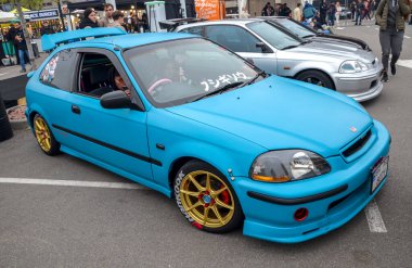 Saldırgan bir şekilde modifiye edilmiş mavi Honda Civic EK hatchback, öne çıkan arka kanat, altın çoklu konuşmalı alaşımlı tekerlekler, kırmızı fren ayarlı ve süspansiyonları indirilmiş.