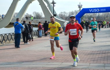 Atletizm sporcuları nehir kenarındaki bir gezinti güvertesinde koşup mavi bir bitiş kemerini geçerler. Şehir arkaplanı ve gülümseyen liderlerle canlı, açık hava yarış sahnesi.