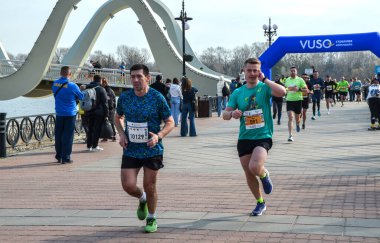 Atletizm sporcuları nehir kenarındaki bir gezinti güvertesinde koşup mavi bir bitiş kemerini geçerler. Şehir arkaplanı ve gülümseyen liderlerle canlı, açık hava yarış sahnesi.