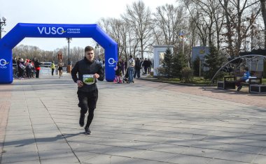 İzleyiciler açık hava yarışını izlerken erkek koşucu geniş bir gezinti alanı boyunca hızla ilerliyor. Mavi şişirilebilir kemer bitiş noktasını gösteriyor, canlı bir atletizm sahnesi oluşturuyor.