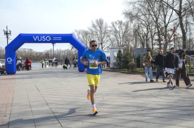 İzleyiciler açık hava yarışını izlerken erkek koşucu geniş bir gezinti alanı boyunca hızla ilerliyor. Mavi şişirilebilir kemer bitiş noktasını gösteriyor, canlı bir atletizm sahnesi oluşturuyor.