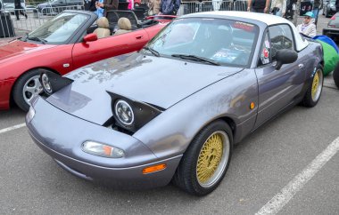 Klasik gri Mazda MX-5 Miata (NA), popüler iki koltuklu roadster, yarı kalkık farları ve göz alıcı altın ağ tekerlekleriyle tanınır.