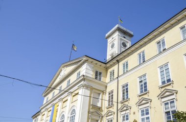 Lviv Belediye Binası 'nın alçak açılı görüntüsü, açık mavi gökyüzüne karşı. Üzerinde bir saat kulesi ve tepeden dalgalanan bir Ukrayna bayrağı var.
