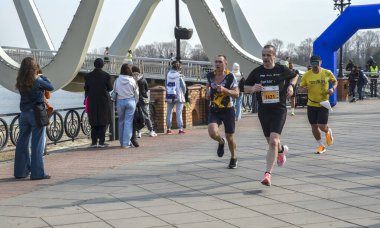 Koşucular nehir kenarındaki bir gezinti güvertesinde koşuyor, seyircilere tezahürat ederken çarpıcı bir modern kemer köprüsü sahneyi çerçeveliyor
