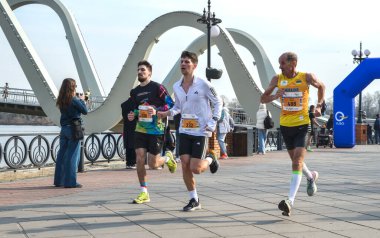 Koşucular nehir kenarındaki bir gezinti güvertesinde koşuyor, seyircilere tezahürat ederken çarpıcı bir modern kemer köprüsü sahneyi çerçeveliyor