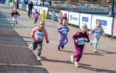 Bir grup gülümseyen kız çocuğu, bir çocuk yarışı sırasında güneşli bir caddede koşar, üzerinde önlükler ve renkli giysiler vardır.
