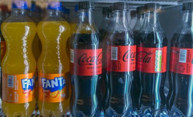 Buzdolabının rafında mavi kapaklı birkaç plastik Fanta Portakal şişesi ve siyah kapaklı Coca-Cola Sıfır Şeker var.