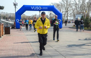 Renkli giyimli koşucular finişe doğru koşuyor. Açık hava parkında, izleyicilerin ve bahar güneşinin olduğu büyük mavi şişme kemerin altında.