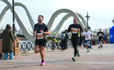 Koşucular nehrin kenarındaki tuğla bir gezinti güvertesinde koşarken, seyirciler yolu izliyor. Sancaklar halka açık bir yarışa işaret ederken, arka planda kemerli bir köprü yükseliyor