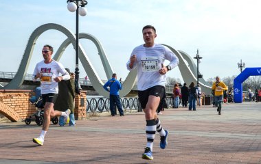Koşucular nehrin kenarındaki tuğla bir gezinti güvertesinde koşarken, seyirciler yolu izliyor. Sancaklar halka açık bir yarışa işaret ederken, arka planda kemerli bir köprü yükseliyor