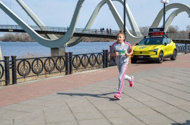 Genç kız modern bir kemer köprüsünün yakınındaki nehir kenarındaki gezinti alanında koşuyor. Zamanlayıcısı olan sarı bir destek aracı sokak yarışını izliyor.