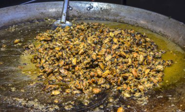 Büyük metal tava lezzetli, yağlı bir sosta pişirilmiş midyelerle doludur. Kırmızı biber ve bitkiler de görülebilir. Deniz ürünleri mutfağı ve küresel sokak yemekleri eğilimleri