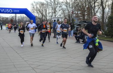 Çeşitli koşucular, mavi şişme kemerin altından geçerek bir meydanda yarışıyor, gündüz sporunda enerji, kararlılık ve toplum ruhunu yakalıyorlar.