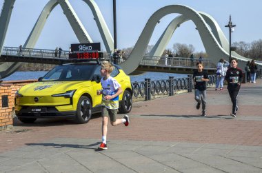 Bir grup genç koşucu nehir kenarındaki gezinti güvertesinde koşuyor ve arka planda belirgin bir kemerli köprü ve tezahürat seyircileri var.