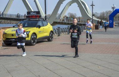 Bir grup genç koşucu nehir kenarındaki gezinti güvertesinde koşuyor ve arka planda belirgin bir kemerli köprü ve tezahürat seyircileri var.