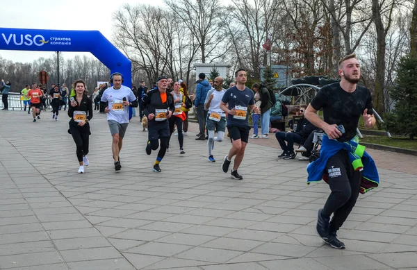 Çeşitli koşucular, mavi şişme kemerin altından geçerek bir meydanda yarışıyor, gündüz sporunda enerji, kararlılık ve toplum ruhunu yakalıyorlar.