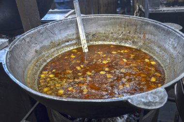 Büyük metal kazan, içinde et ve sebze parçaları bulunan kırmızı-kahverengi bir çorbayla dolu bir ateşin üzerinde oturur. Tencerenin içinde uzun metal bir kepçe var.