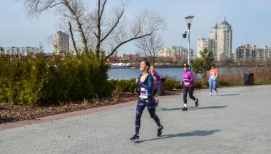Atletik teçhizatlı bir grup kız koşucu şehrin ufuk çizgisi boyunca asfalt yol boyunca koşuyor. Parlak gün, yeşillik ve şehir enerjisi aktif bir açık hava sahnesi oluşturur