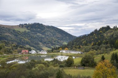 Küçük bir köy durgun bir göletin kenarında, beyaz ve kırmızı çatılı binaları yansıtıyor. Yuvarlanan, ormanlık tepeler bulutlu bir gökyüzünün altında yükseliyor, ağaçlardaki sonbahar renklerinin dokunuşlarıyla