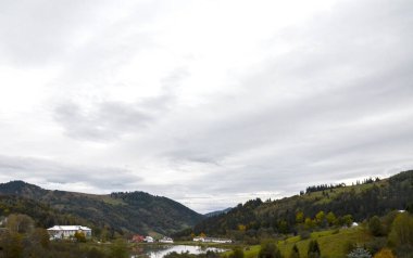 Küçük bir köy durgun bir göletin kenarında, beyaz ve kırmızı çatılı binaları yansıtıyor. Yuvarlanan, ormanlık tepeler bulutlu bir gökyüzünün altında yükseliyor, ağaçlardaki sonbahar renklerinin dokunuşlarıyla