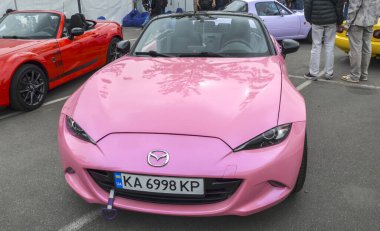 Üstü açık, modern pembe Mazda Miata. Araç, benzersiz rengini ve ikonik spor araba siluetini vurgulayarak halka açık bir etkinlikte sergileniyor.