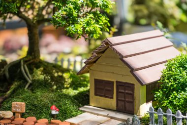 Küçük bir ev, ağaç, heykelcik ve yeşillik ve bitkilerle çevrili dekoratif bir yolun yer aldığı bir tencerede minyatür bahçe manzarası.