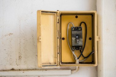 Beyaz bir duvara monte edilmiş eski sarı elektrik düğmesi kutusu. Panelin içinde görünür kablolar ve toz birikiyor.