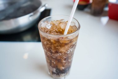 Buzlu kolayla dolu bir bardağın yakın çekimi ve restoran ortamında beyaz bir masanın üstünde pipet.