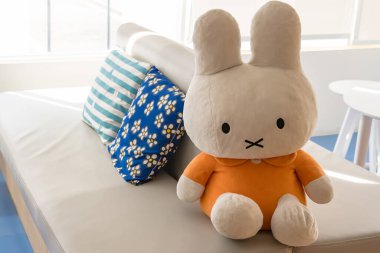 Büyük peluş, turuncu elbiseli tavşan oyuncağı. Kapalı bir mekanda güneşli bir pencerenin yanında, sandalyede oturuyor.