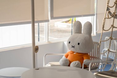 Büyük peluş, turuncu elbiseli tavşan oyuncağı. Kapalı bir mekanda güneşli bir pencerenin yanında, sandalyede oturuyor.